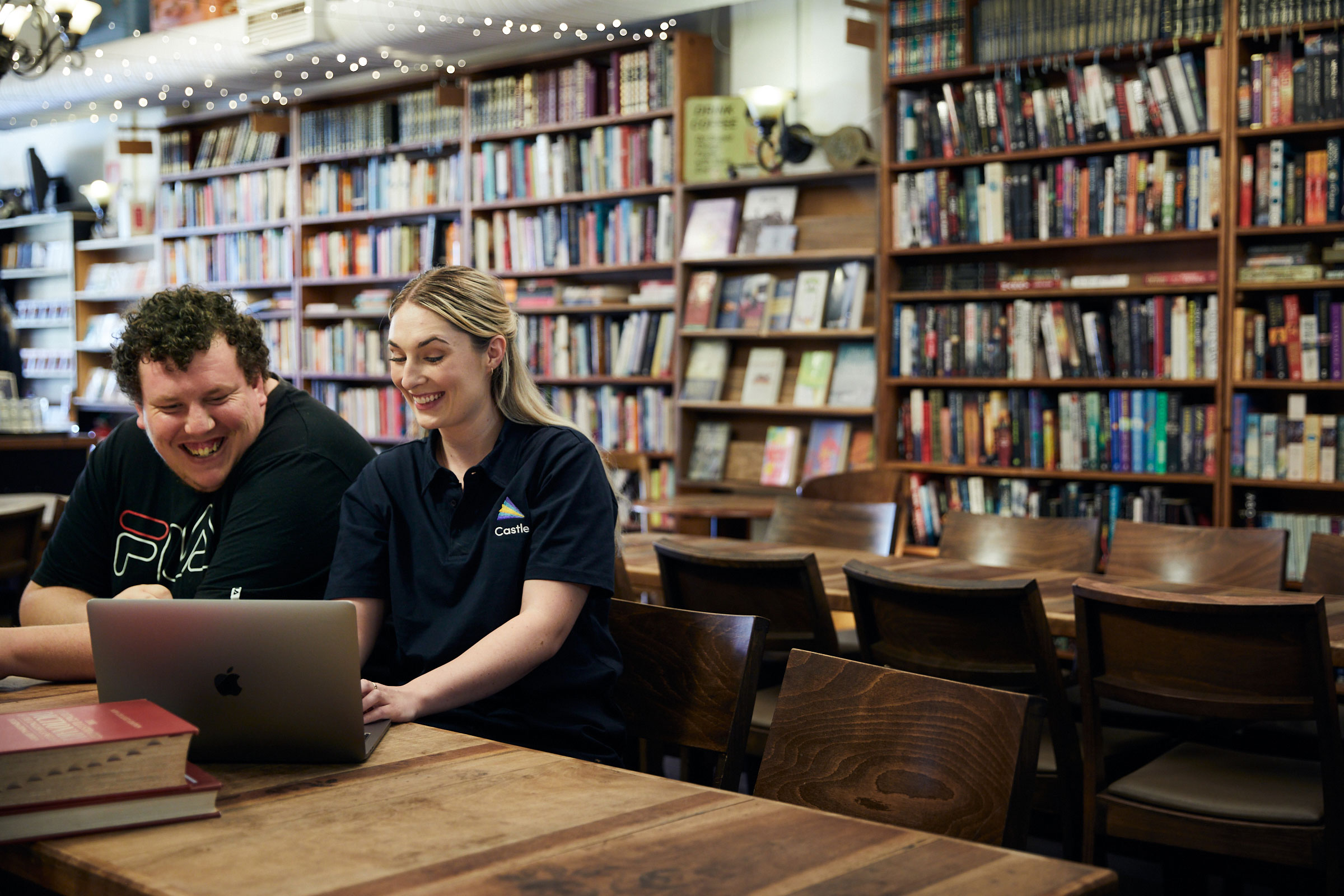 participant and Castle support worker using a laptop together, sitting in front of a long bookcase.